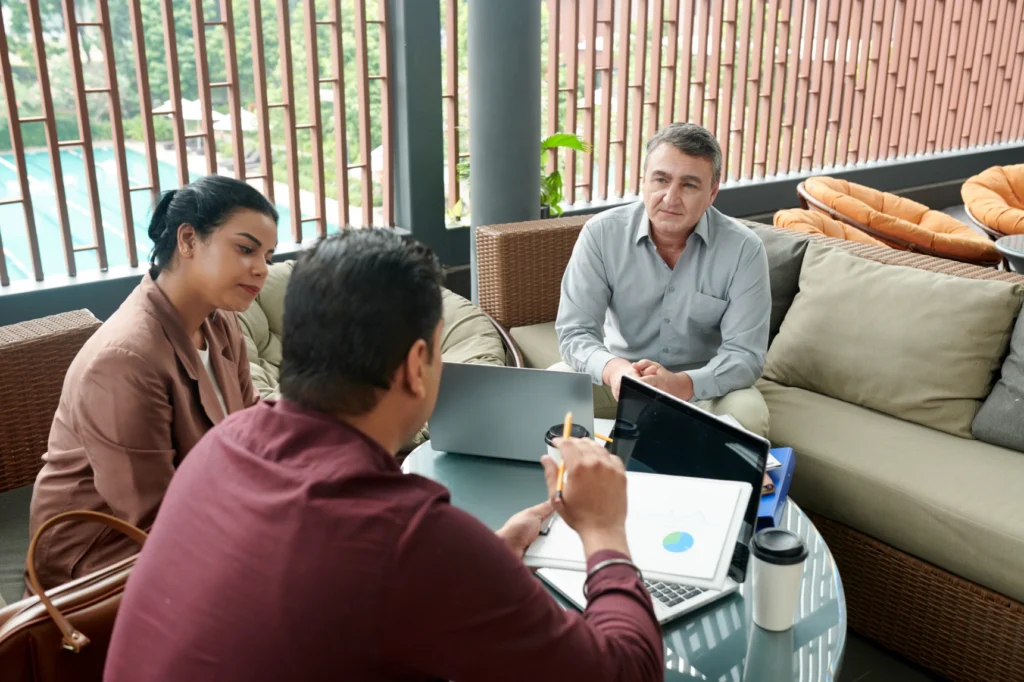 Three professionals in a meeting, discussing over a laptop in a casual outdoor setting.